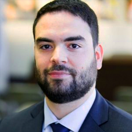 Man with dark hair and beard wearing a suit and tie, looking at the viewer.