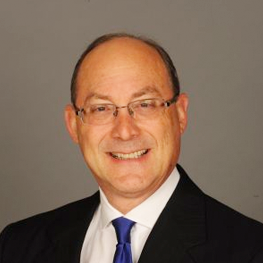 Man wearing glasses, blue tie, and black suit smiling.