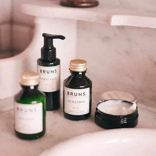 Op een marmeren oppervlak staan ​​vier donkere glazen flessen en een klein open potje met haar- of huidverzorgingsproducten van het merk Bruns.