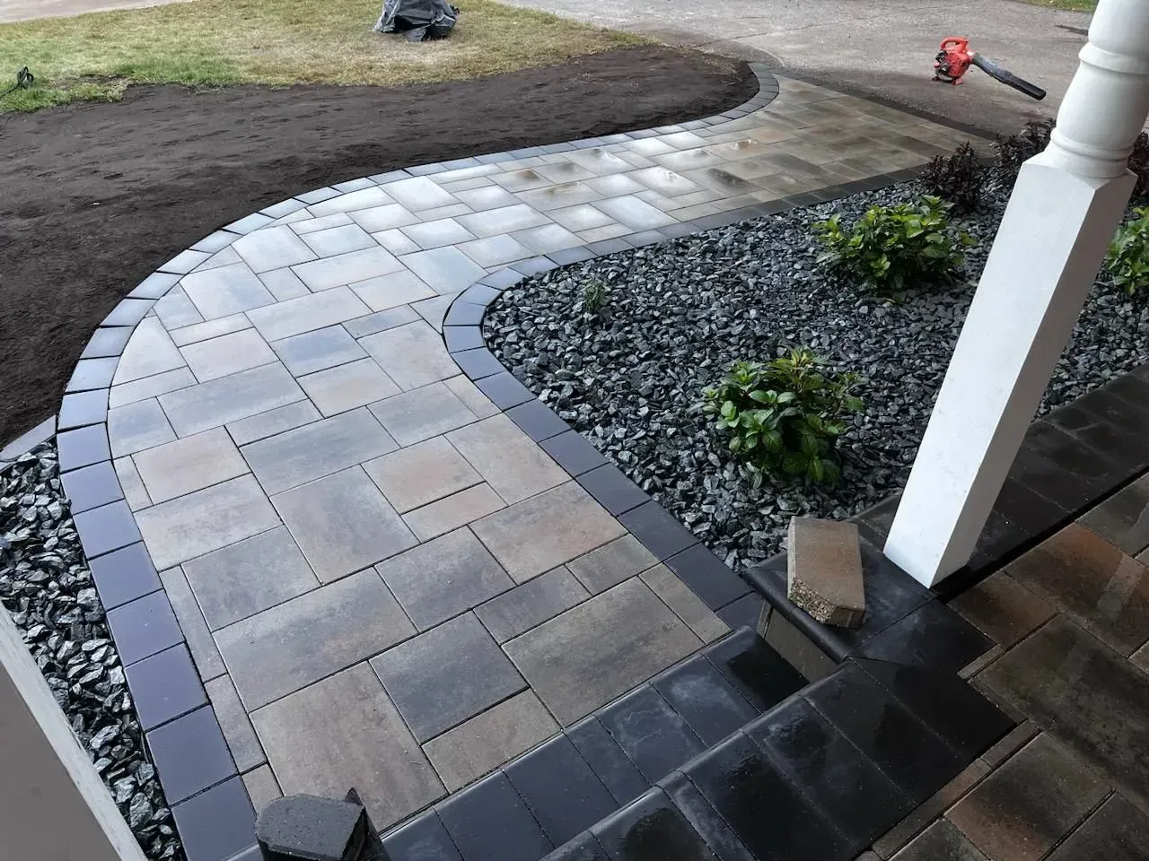 A brick walkway with a black border curves from the dark gray mulch-covered landscaping towards the entrance stairs.