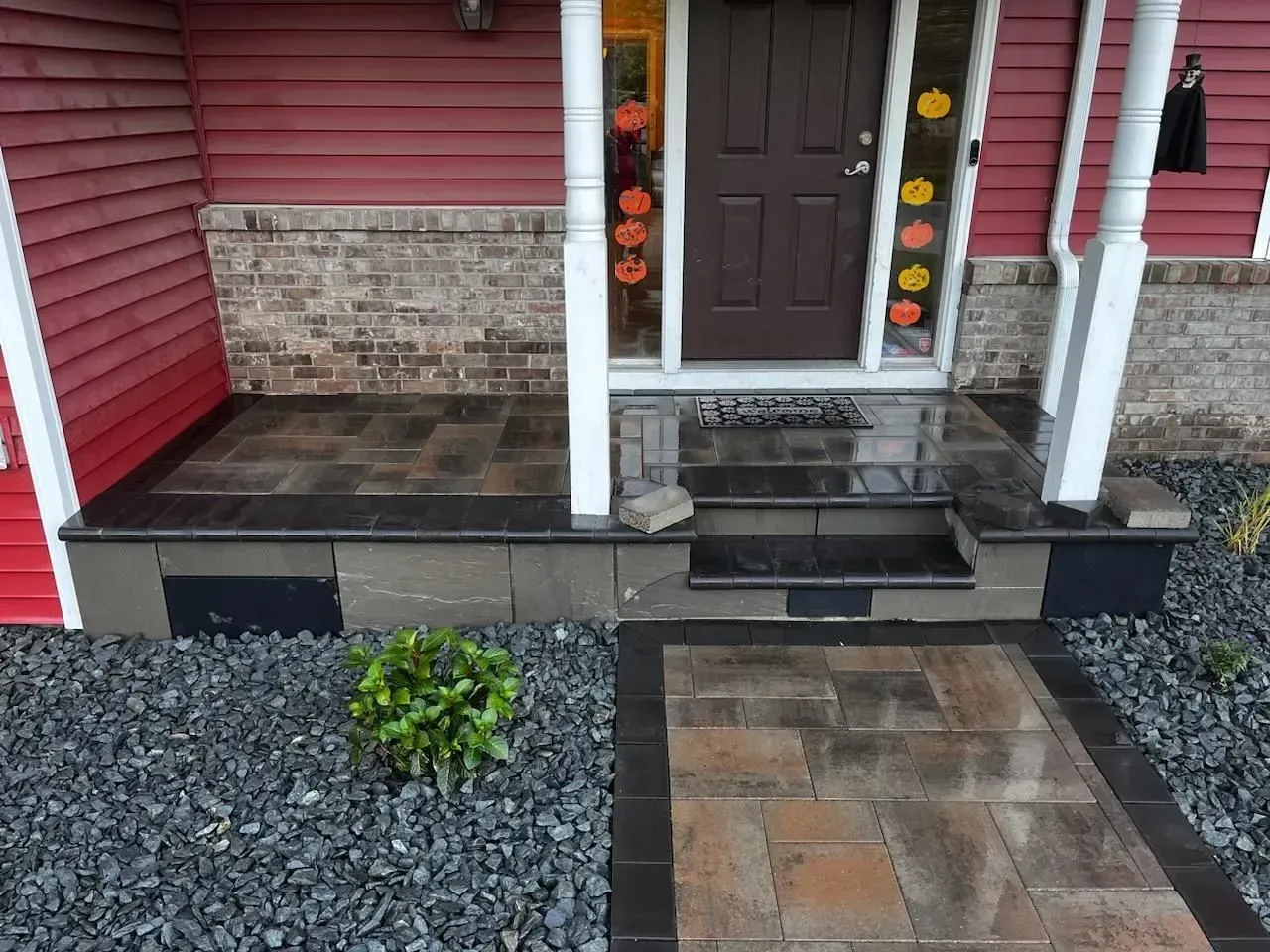 A front porch with dark pavers, steps, and a walkway leading to a brown door.  Black stone borders the walkway and steps.