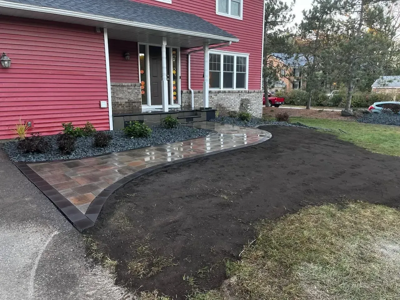 Red house with a newly paved walkway leading to the front door; landscaping is being done on the lawn.