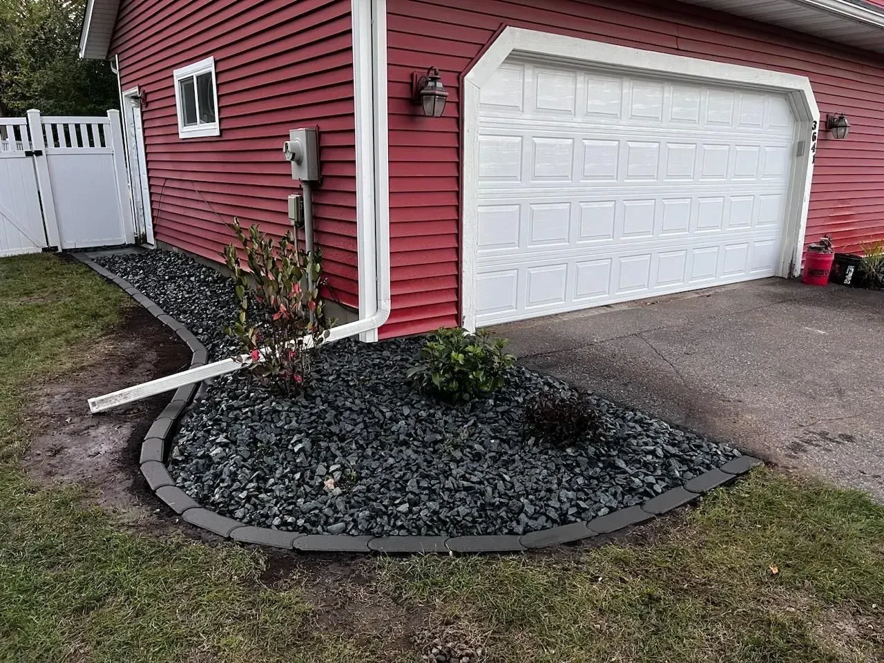 Red-sided garage with white garage door, bordered by a black stone garden bed edging the grass.