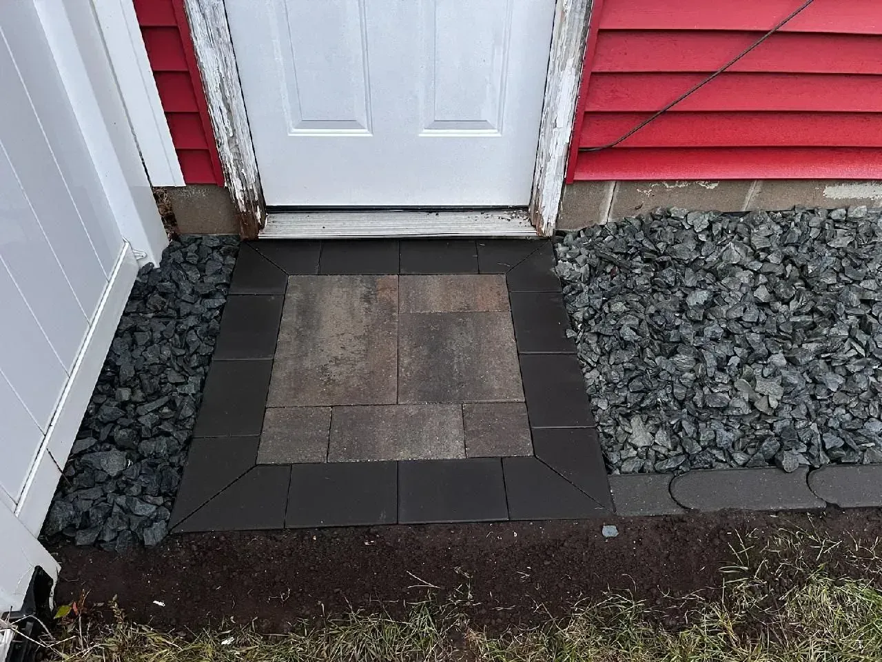 A small brick patio with a dark border and a lighter center, in front of a white door. Dark stones surround it.
