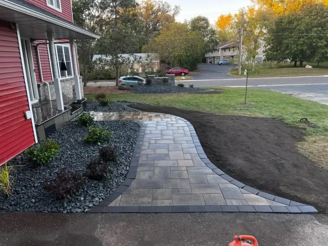 A paved pathway curves from a red house to the street. Landscaping features dark rocks and plants.