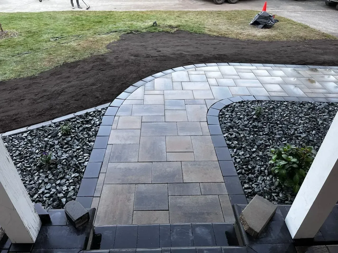 Paver pathway curving to a porch, surrounded by gravel, grass, and newly laid soil.