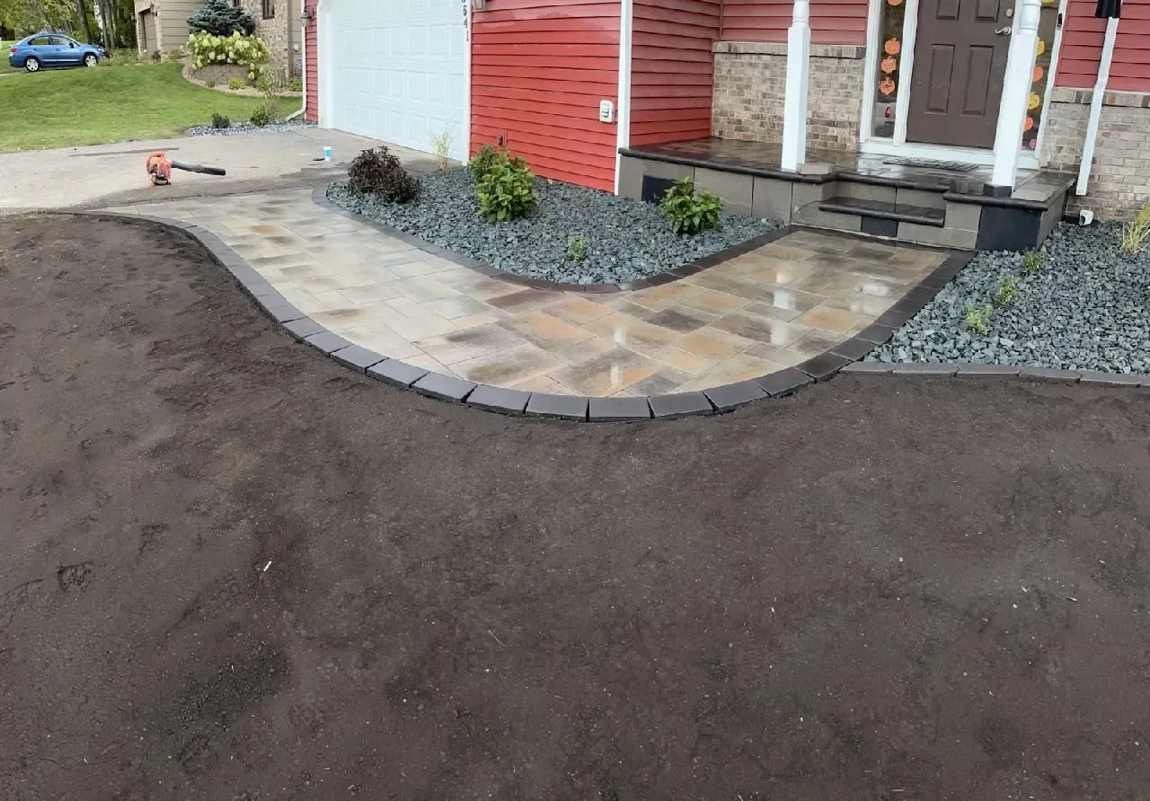 A paved walkway leads to a home's entrance. The path has a brick border and borders a dark gravel bed with bushes.