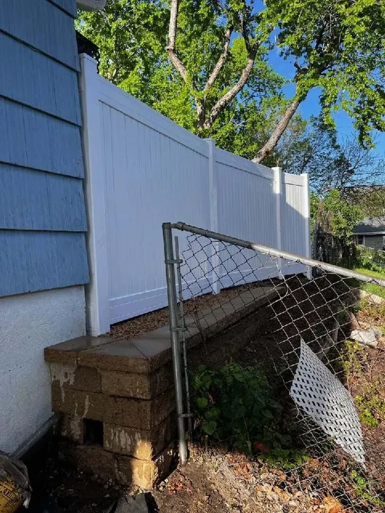White vinyl fence next to a light blue house with a crumbling brick foundation. A bent chain link fence is in the foreground.