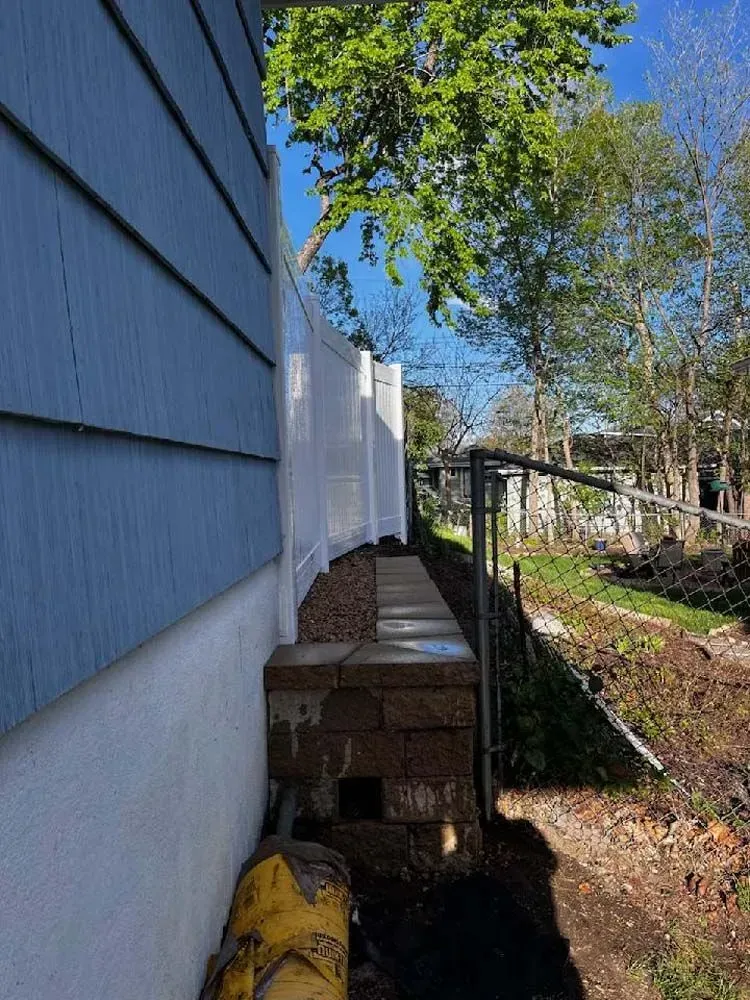 Blue siding next to a retaining wall and white fence. Yellow pipe at the bottom. Trees and dirt in the background.