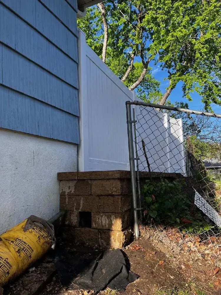 Side view of a house with blue siding, a white fence, and a chain-link gate; stacked concrete blocks are at the base 