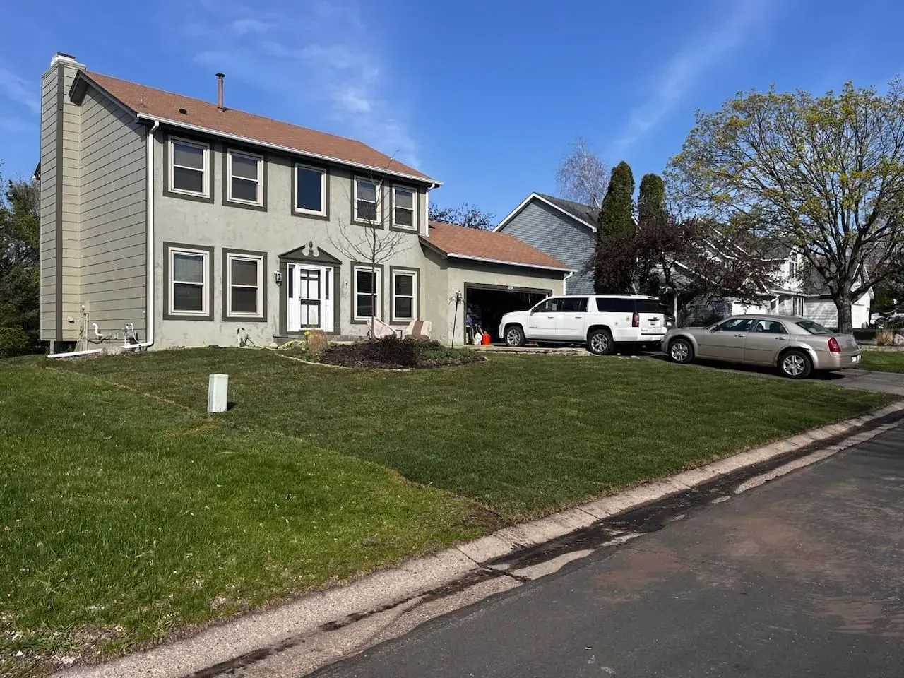 Two-story house with tan siding, green lawn, driveway, and two vehicles parked in front, under a clear blue sky.