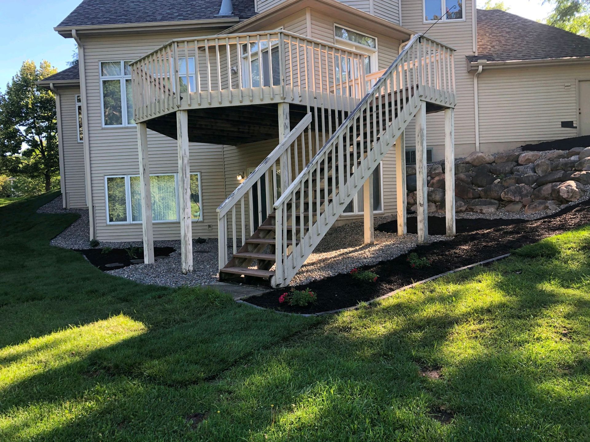 Wooden deck with stairs attached to a two-story beige house, with landscaping and a grassy lawn.