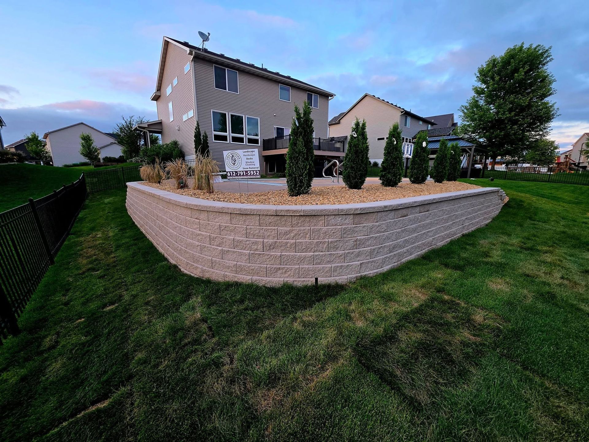 A backyard with a two-story house, a curved retaining wall, and evergreen trees. The lawn is green under a cloudy sky.