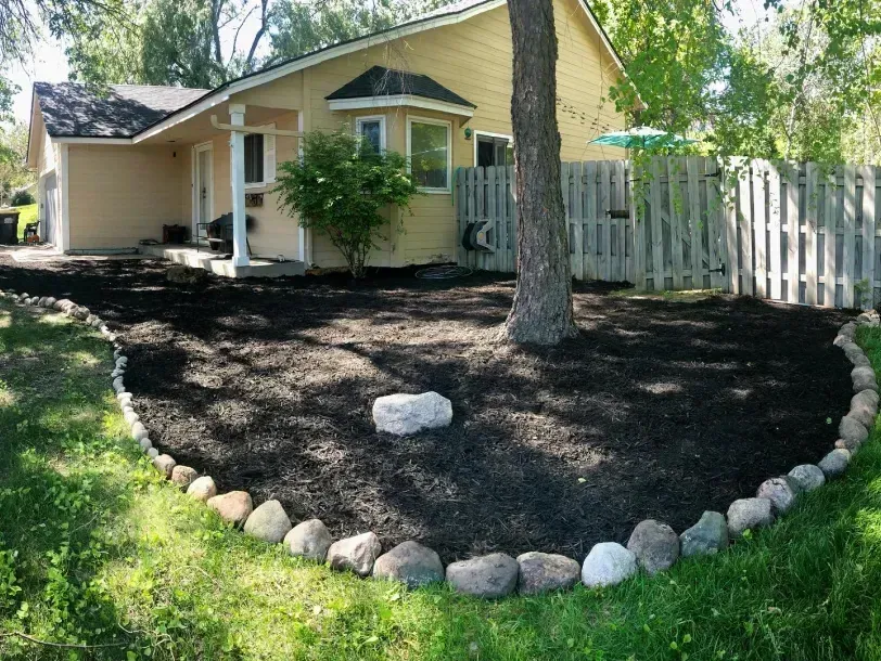 A house with a mulched garden bed bordered by stones, wrapping around a tree. The house is yellow with a dark roof.