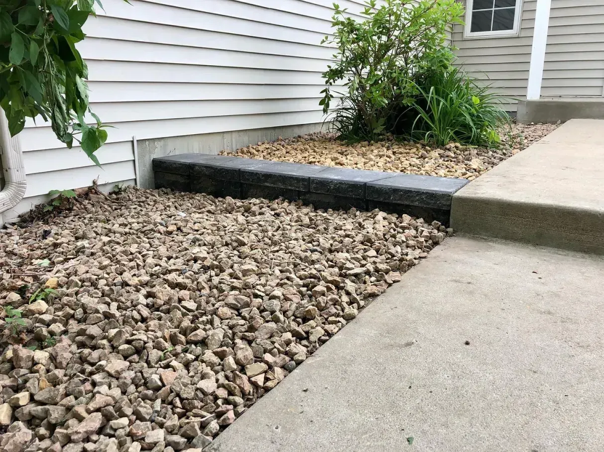 Gravel landscaping bed with a concrete edge next to a sidewalk and house. A few plants are visible in the bed.