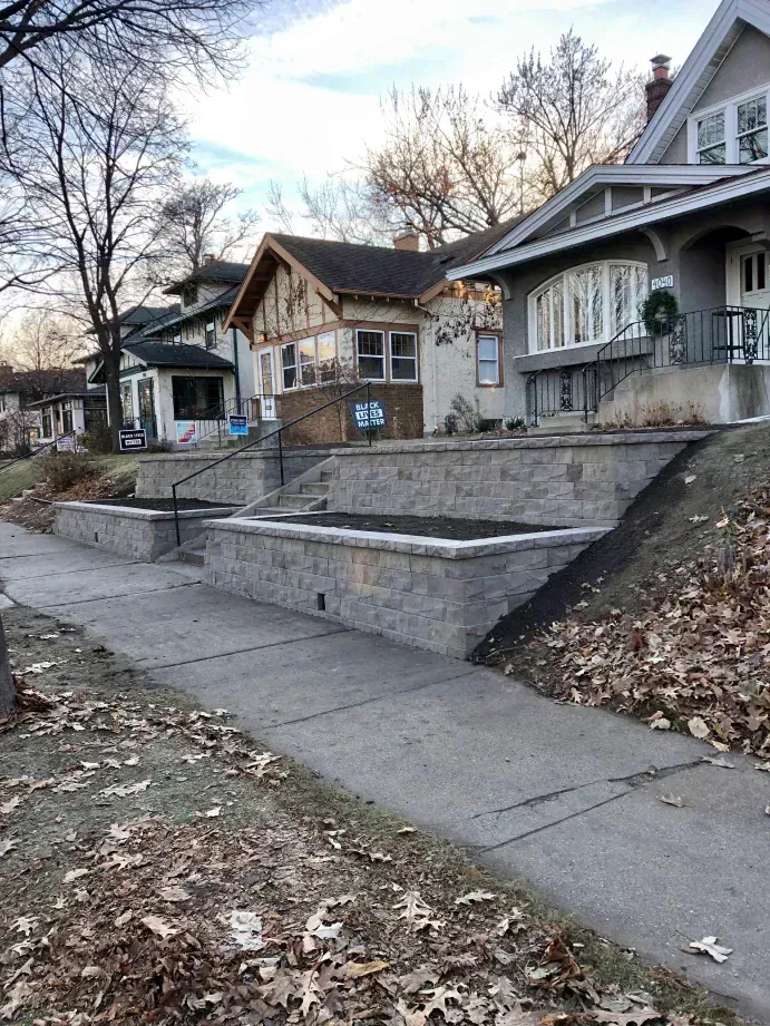 Sidewalk in front of several houses, with a tiered retaining wall along the sidewalk. Fallen leaves are on the ground.