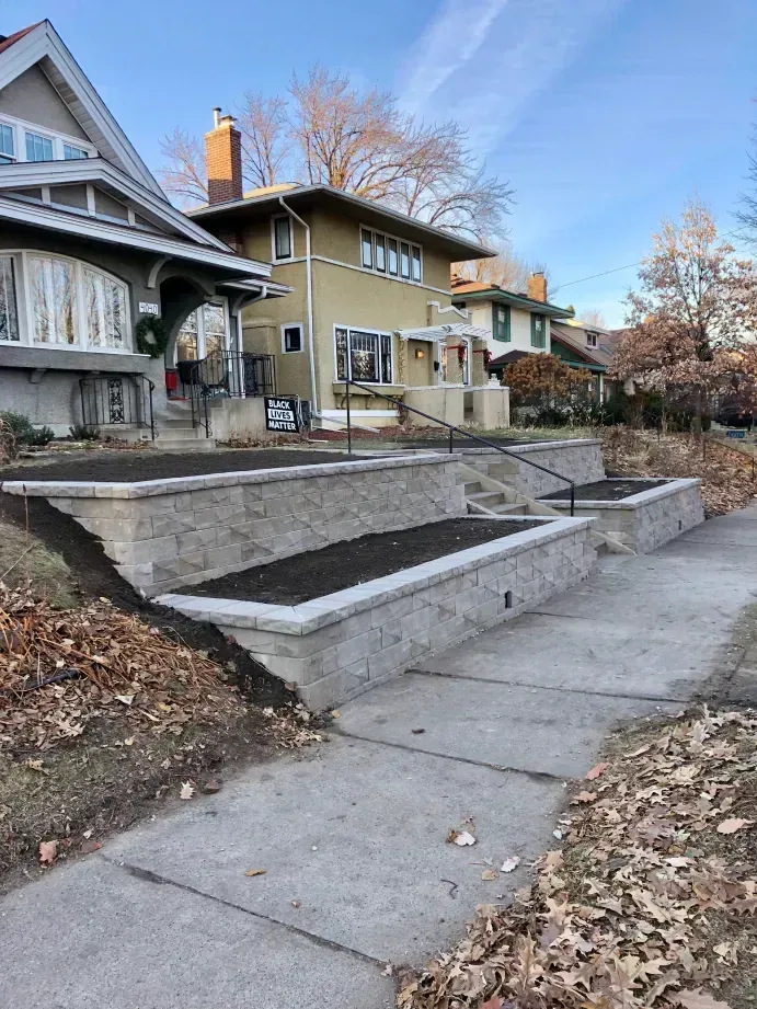 Stone retaining walls with planted beds step down from a sidewalk, leading to residential houses on a fall day.