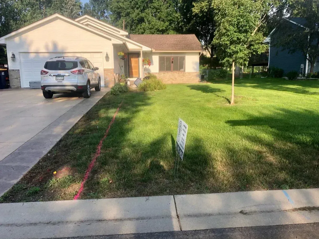 A beige house with a white SUV parked in the driveway. A sign sits in the front yard, which is mostly green grass.