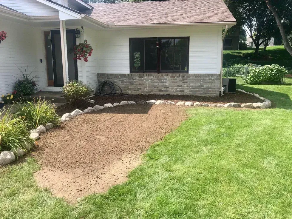 A small patch of freshly tilled dirt surrounded by rocks and grass sits in front of a white house with a brown roof.