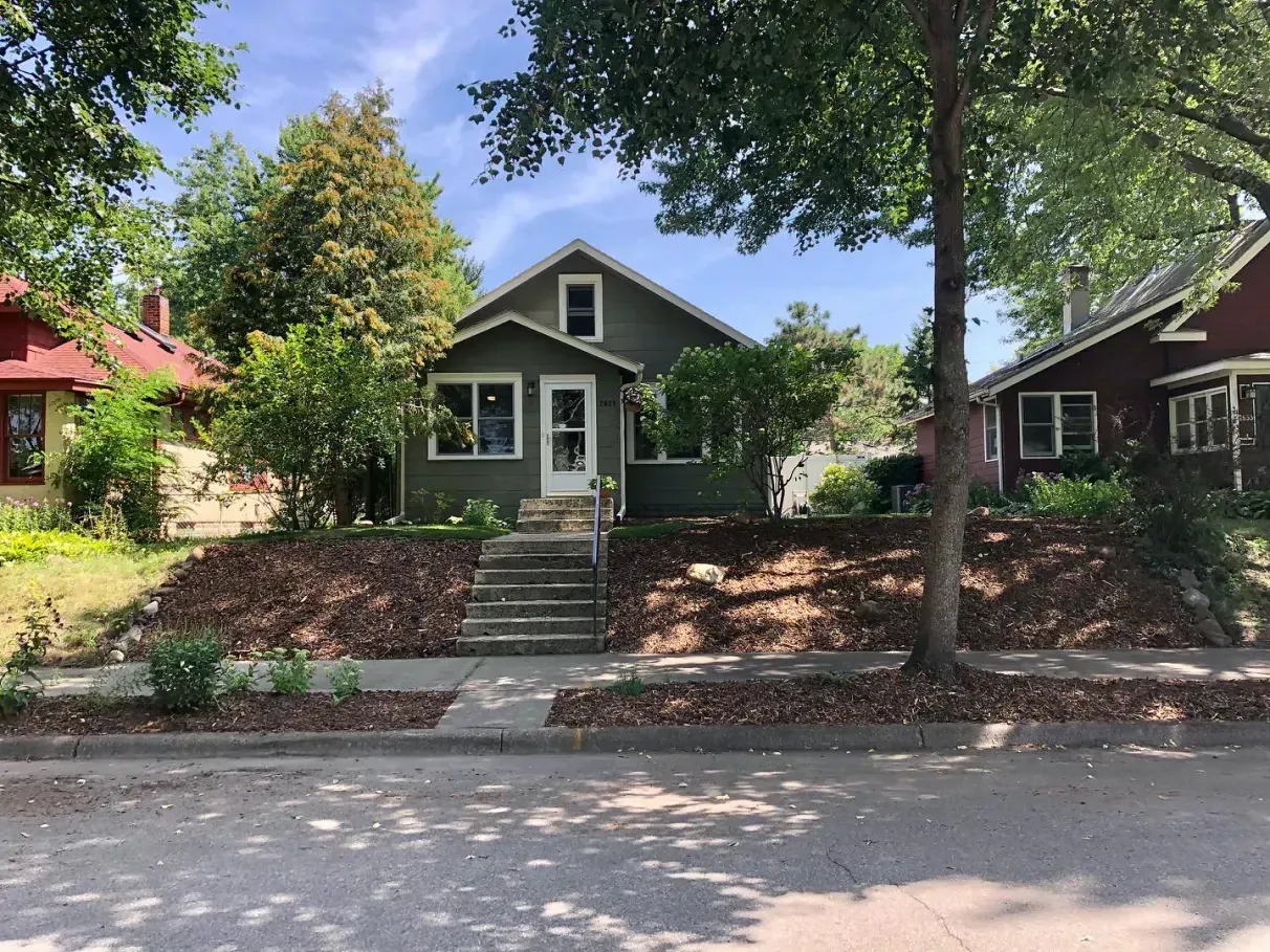 Green house with a small porch and steps leading up from the sidewalk. Trees and neighboring houses are visible.