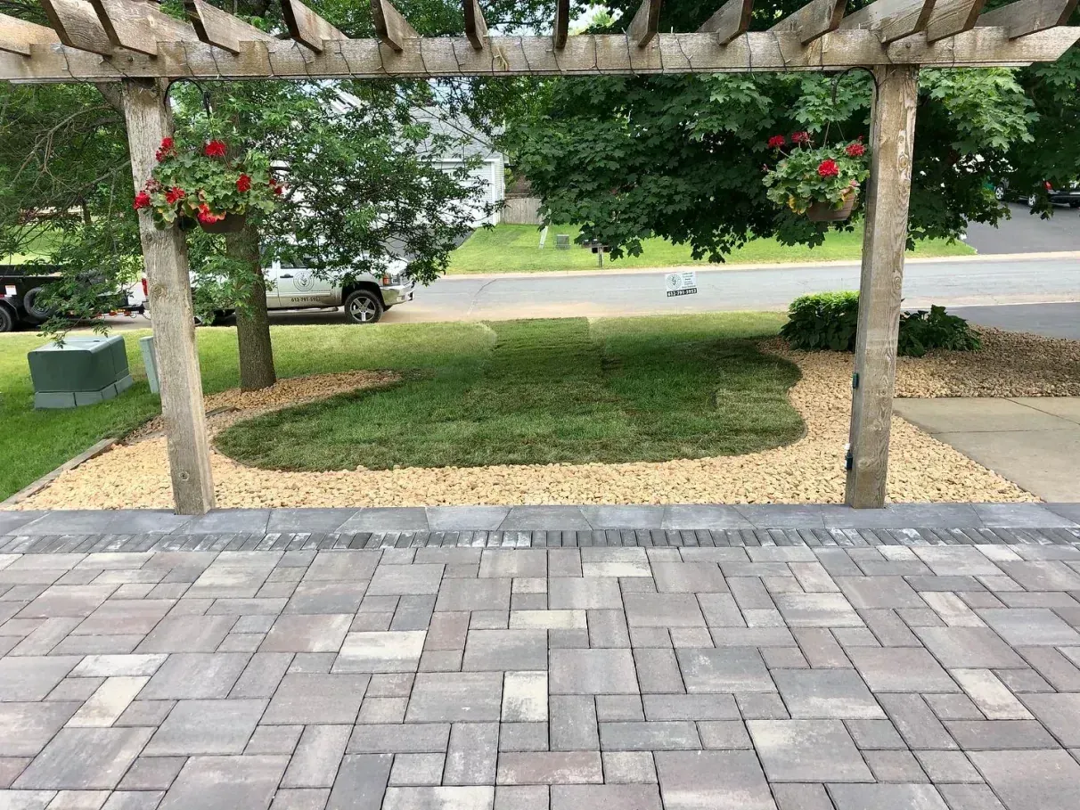 A landscaped outdoor space featuring a brick patio, gravel, grass, and a wooden pergola with hanging flower baskets.