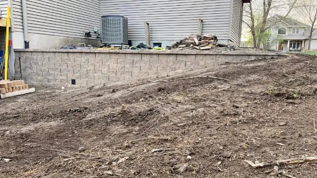A retaining wall made of stone bricks supporting a yard next to a house. The yard is dirt, & there's an AC unit on the wall.