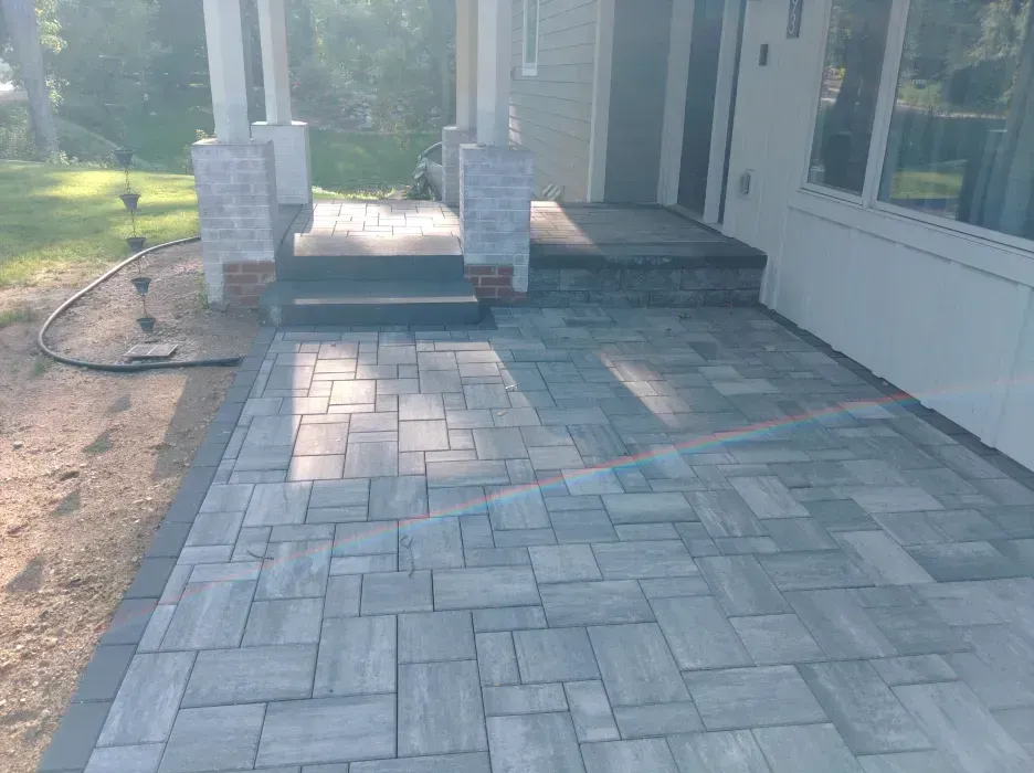 A newly constructed gray brick paver patio and porch. The porch has white columns & steps. Sunlight is cast across the scene.