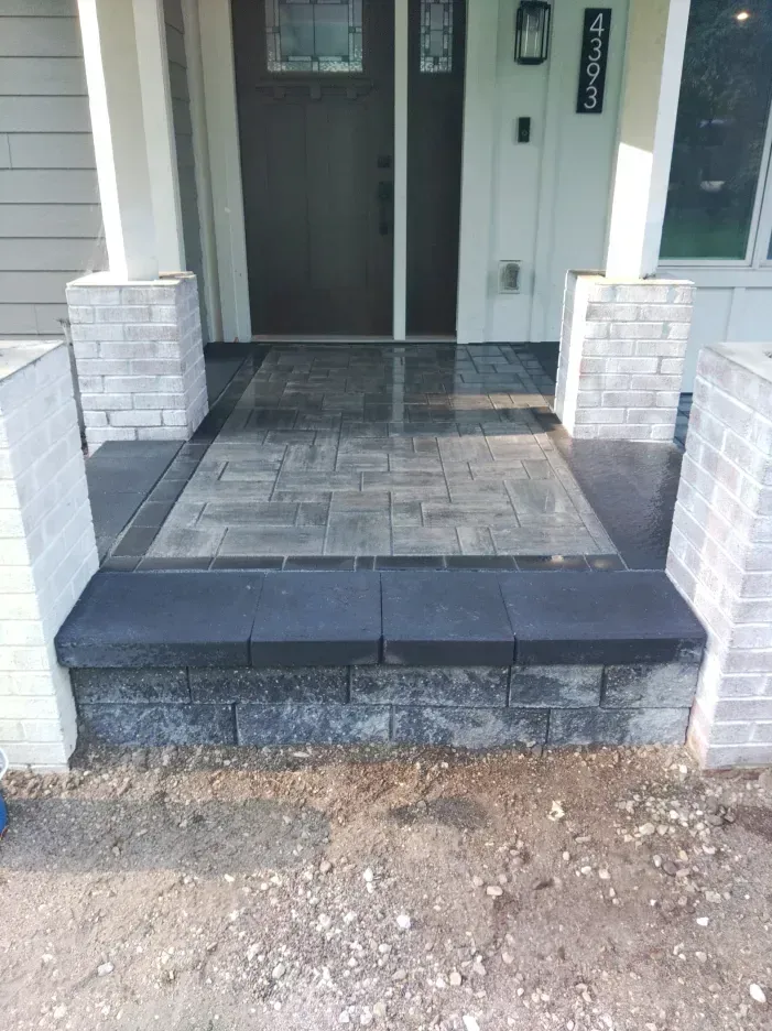 A front porch with a gray brick step and textured pavers leading to a dark door, flanked by white columns.