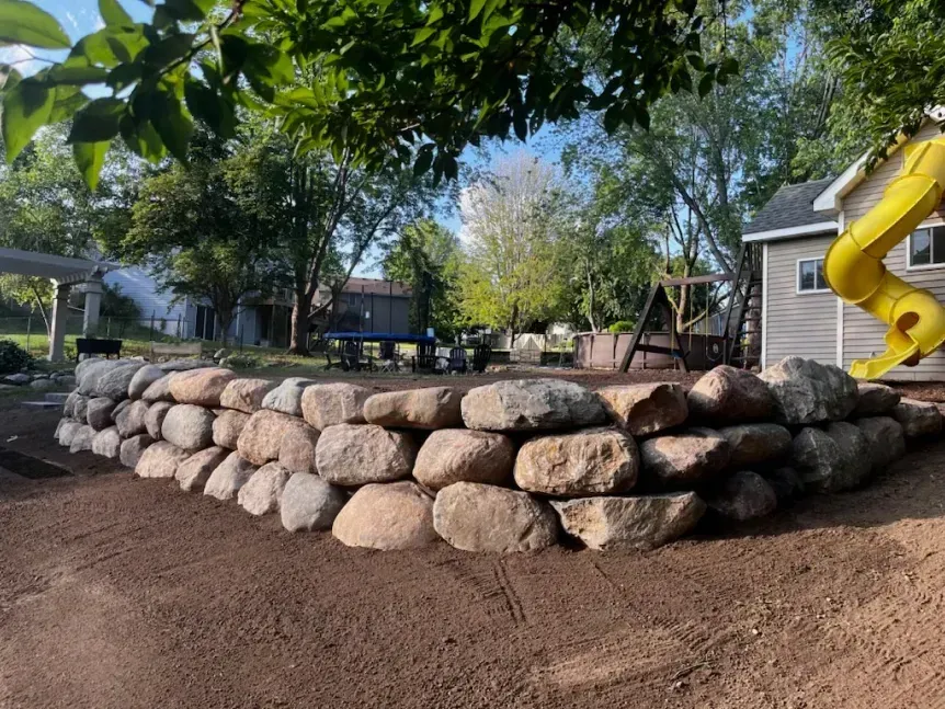 Rock retaining wall in a yard with a playground in the background. Yellow slide on the right, blue table.
