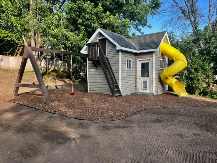 Playhouse with a yellow slide and a wooden swing set on a wood-chip ground.  Stairs lead up to the playhouse balcony.