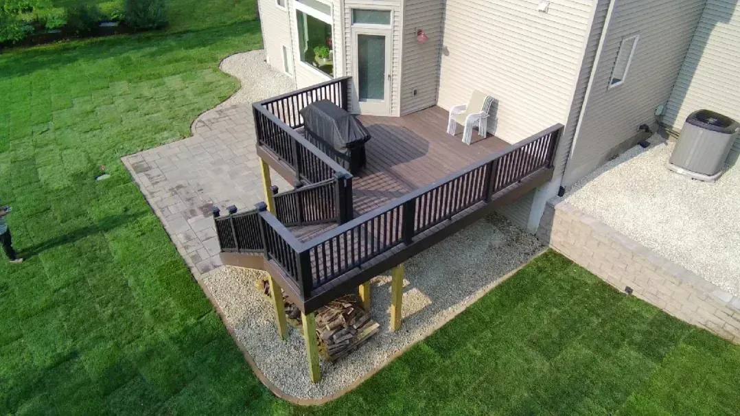 An aerial view of a deck attached to a house with a grill and a small chair; surrounded by a gravel patio and green grass.