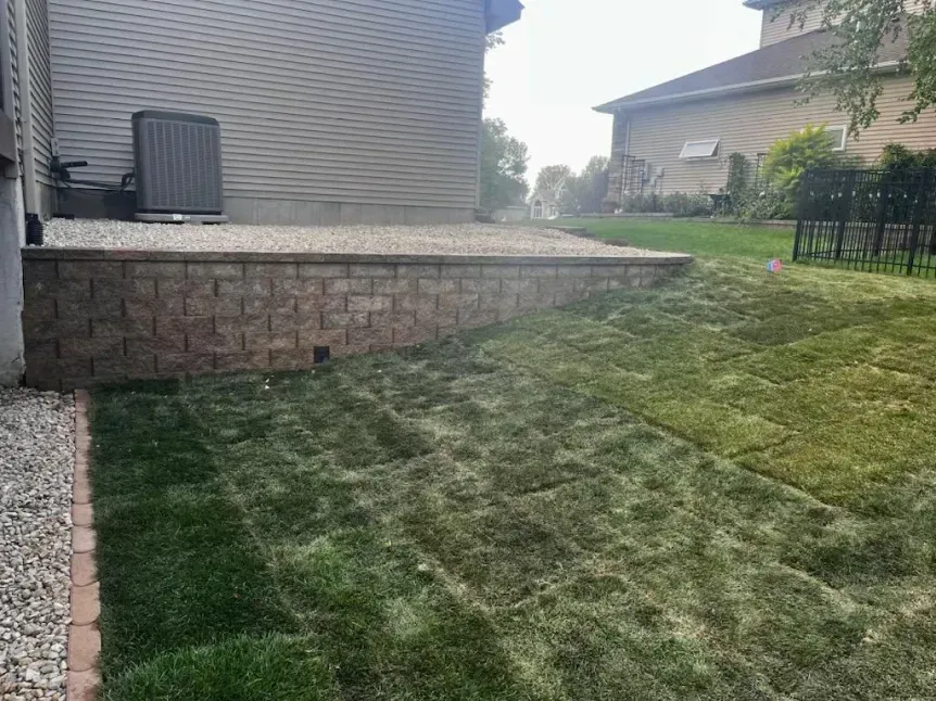 A retaining wall made of brown blocks supporting a patio next to a house, with newly laid grass in the foreground.