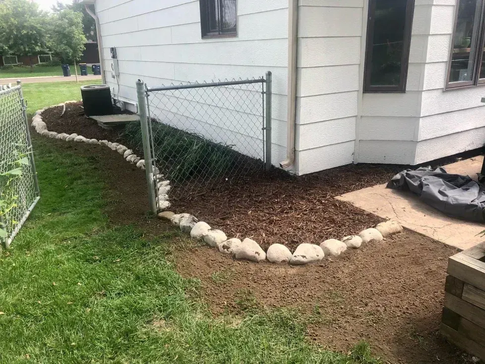 A house corner with a garden bed edged with stones. The bed is covered in mulch and bordered by a chain-link fence.