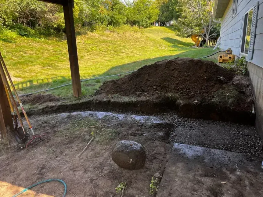 Construction site with a dirt pile, trench, and exposed foundation next to a house and grassy hill.