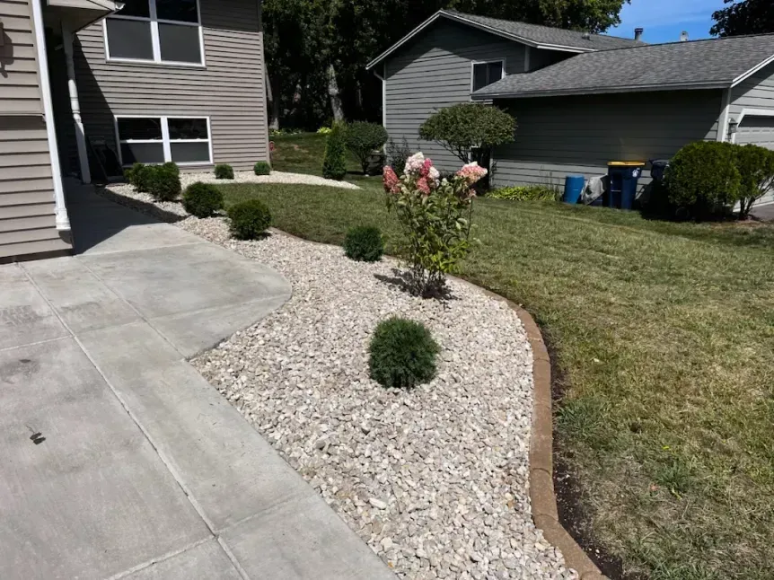 Concrete sidewalk leads to a house with a landscaped rock garden, small green shrubs, and a flowering tree.