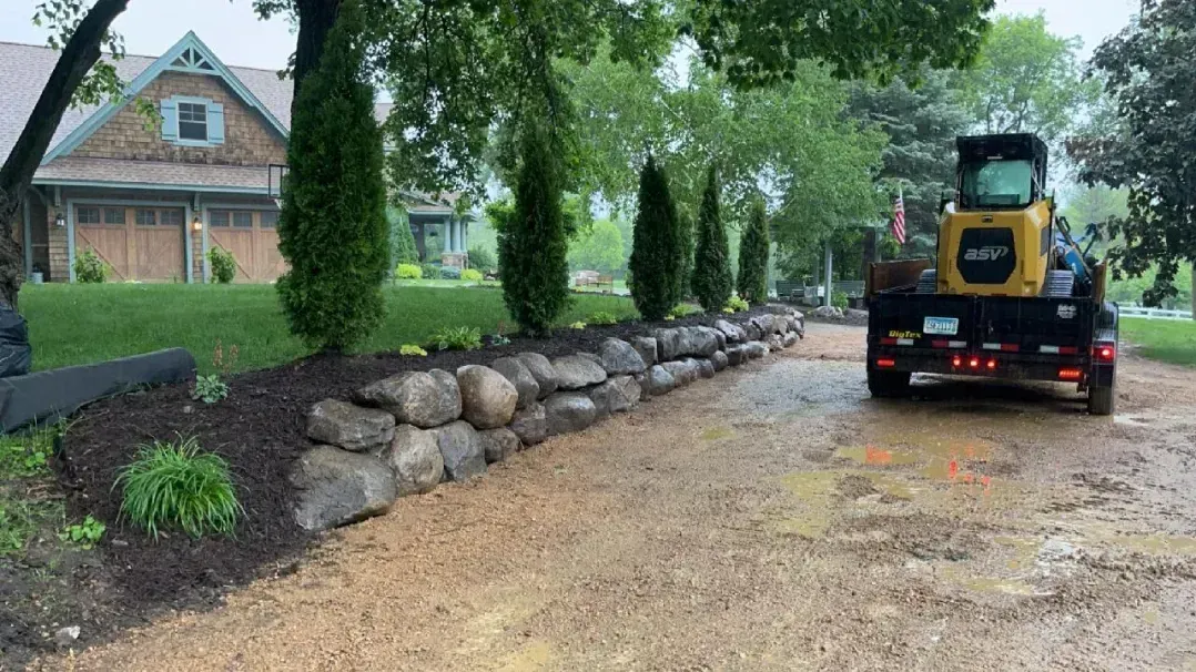 A muddy driveway with a trailer carrying a small construction vehicle, next to a stone retaining wall in front of a house.