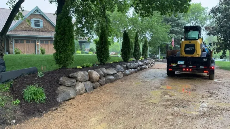 A yellow construction vehicle on a flatbed trailer parked on a muddy driveway next to a stone retaining wall and a house.