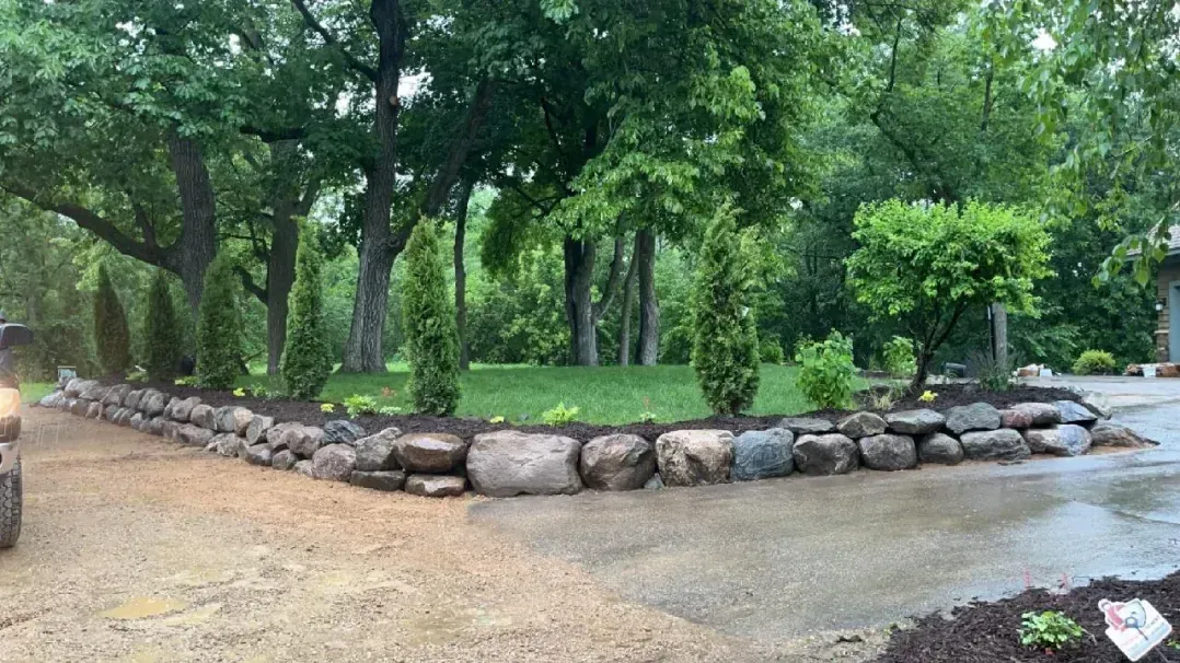 A stone retaining wall frames a green lawn and young trees. A gravel driveway curves around the landscaped area.