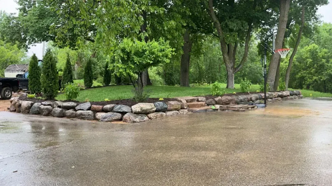 A wet driveway with a rock retaining wall and recently planted trees, with a lush green lawn in the background.