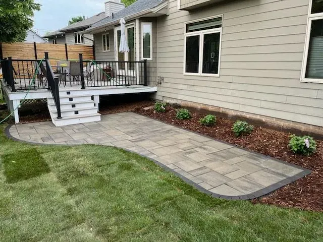 A paved patio and deck with steps leading to a backyard. Light brown pavers are surrounded by grass and landscaping.