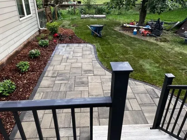 Paver patio and walkway lead from a house with black railings to a grass yard, landscaped with shrubs and mulch.