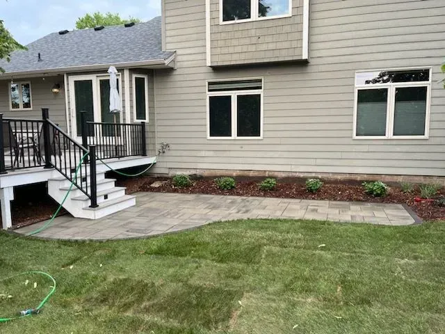 Backyard patio with a paver path, deck, and house. Green grass surrounds the patio and flower bed.