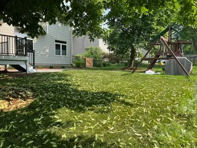 Backyard with grass covered in debris, a swing set, a deck, and a two-story house under a partly cloudy sky.