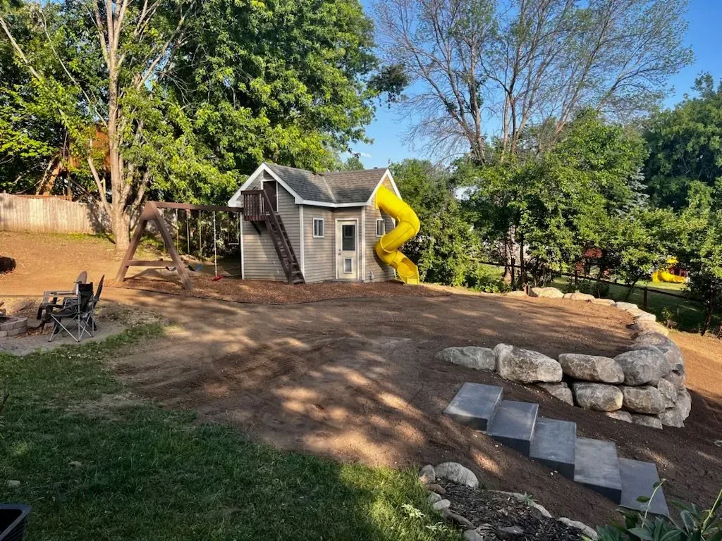 A backyard playset with a gray house, yellow slide, swings, and a stone retaining wall with stairs.