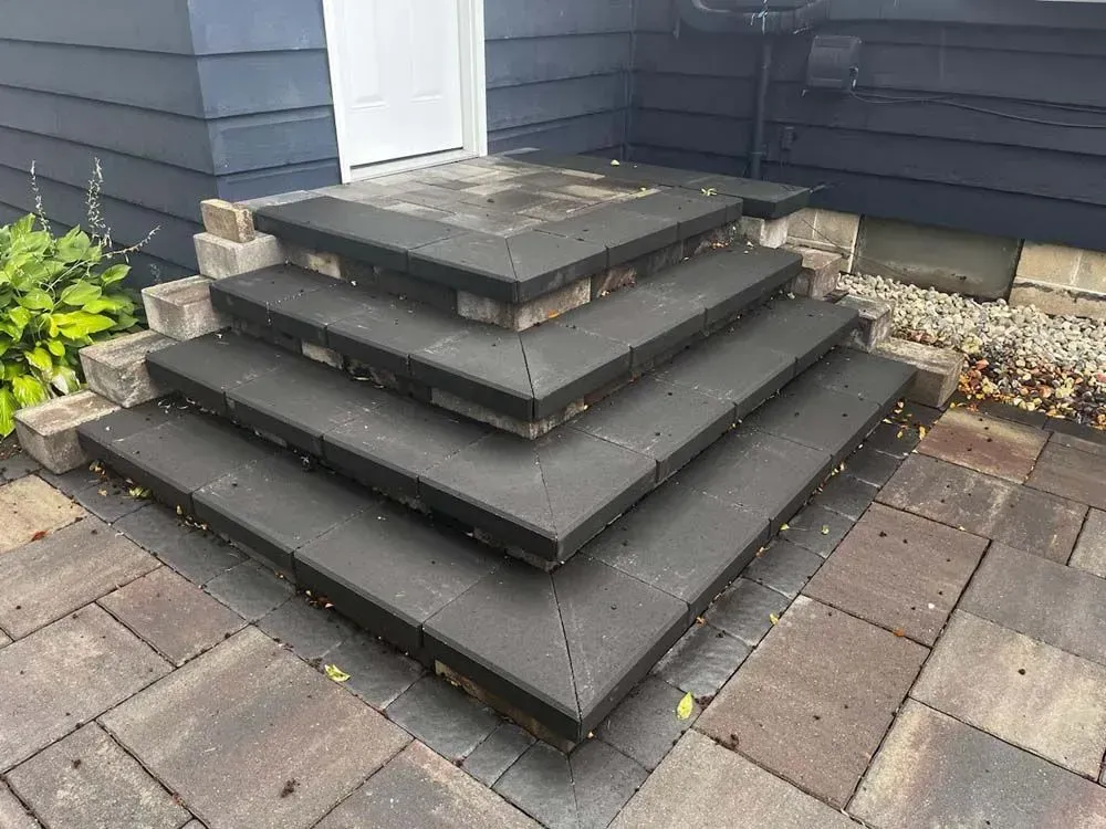 Brick steps leading to a doorway with dark pavers and a brick border. Grey siding on the building to the left.