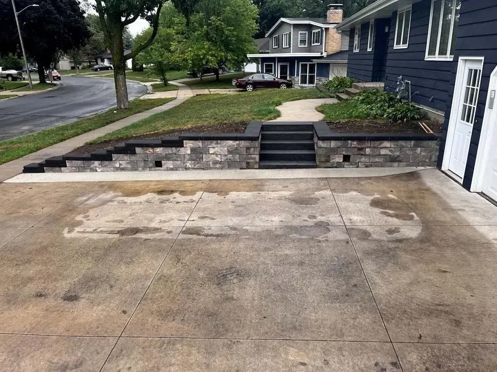 Gray brick retaining wall and steps lead to a grassy area. A driveway is in front of 
