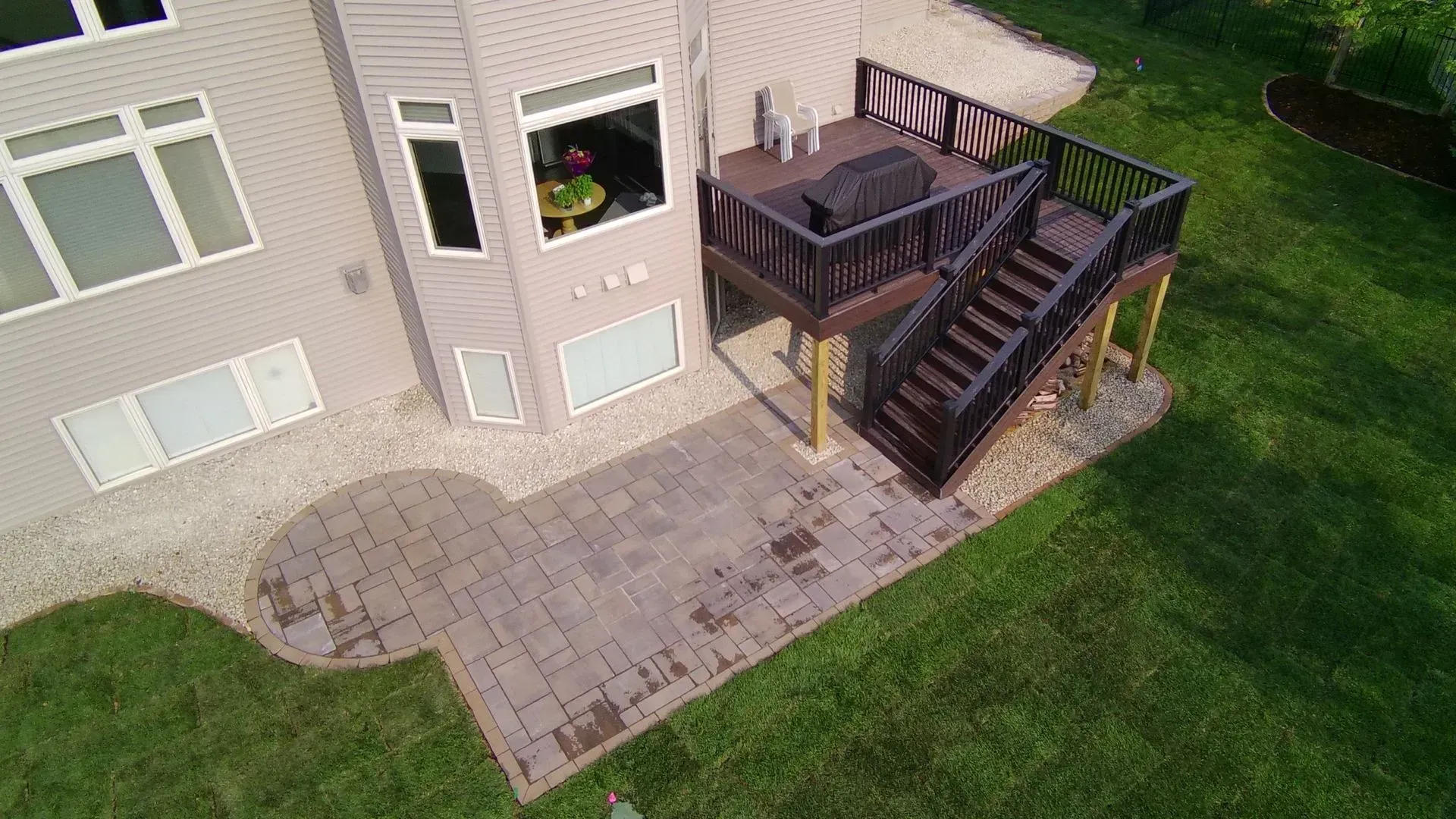 High-angle view of a house with a deck and paved patio. A dark brown deck has stairs leading to the ground.