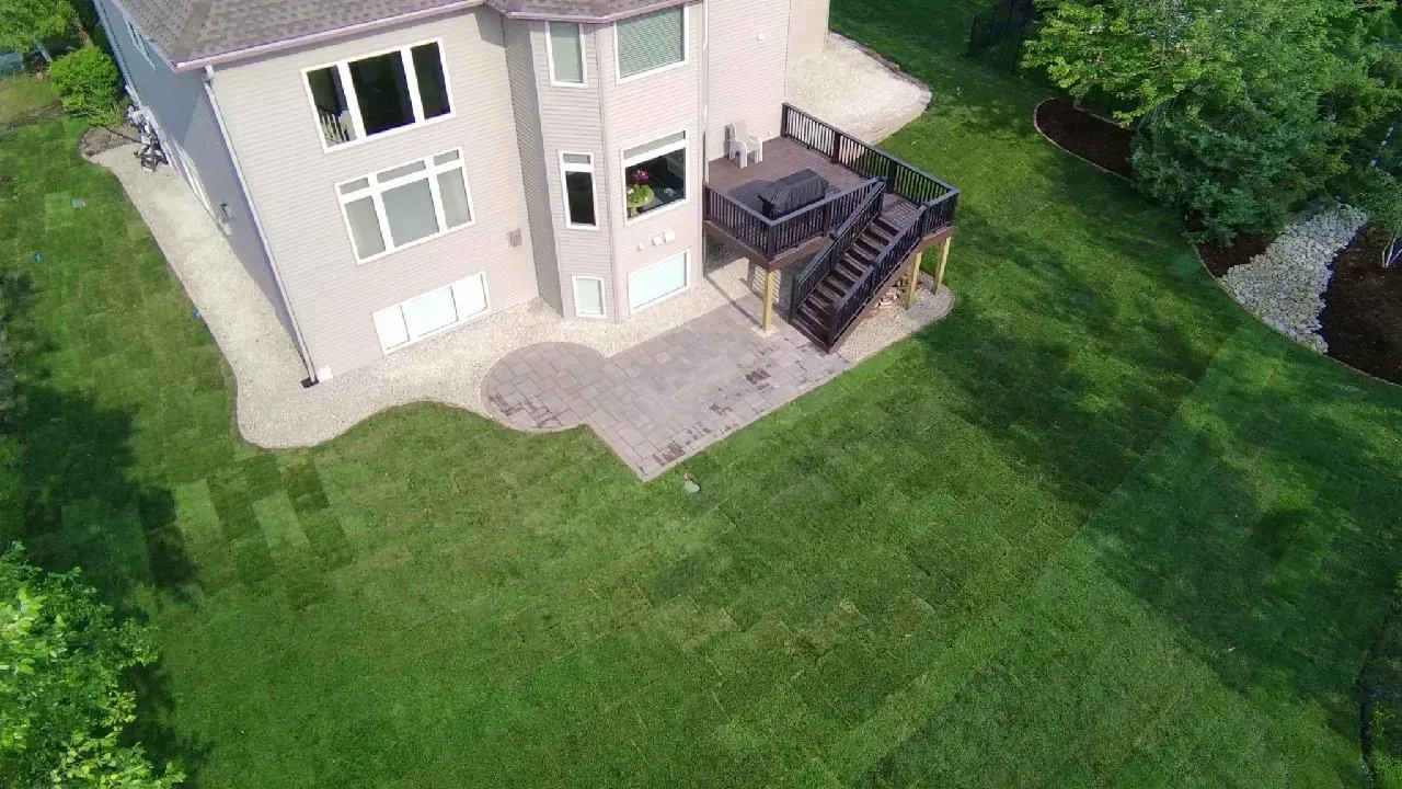 An aerial view of a beige house with a dark deck and patio, surrounded by a green lawn.