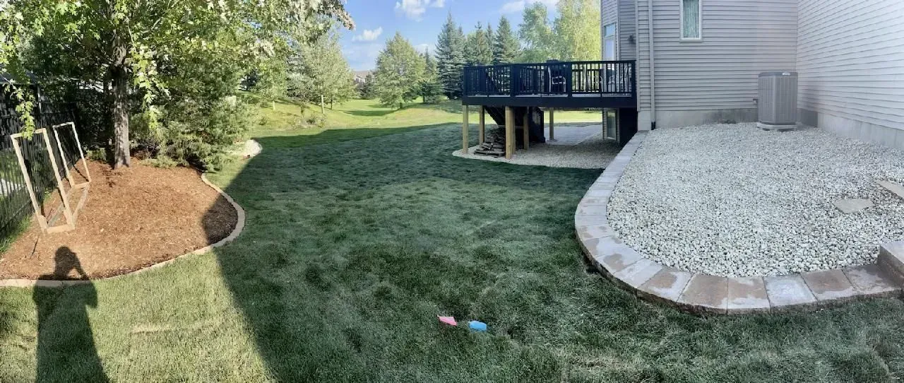 A backyard with green grass, a wooden deck, and a gravel patio.  There is a swing set and some mulch beds.