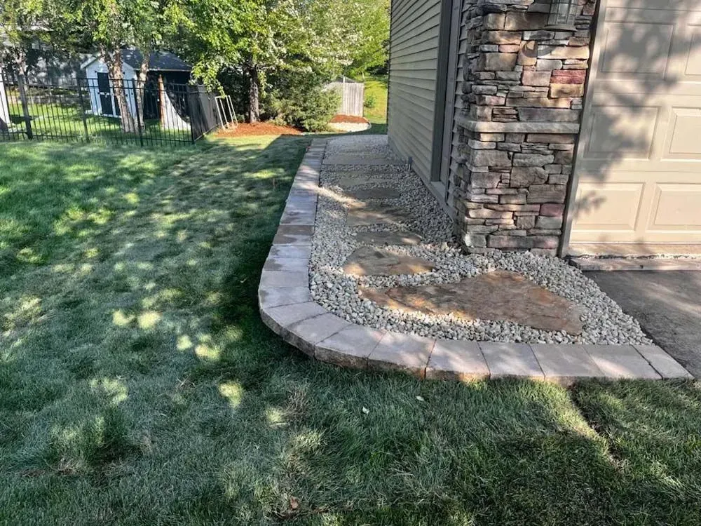 A house corner with stone veneer next to a concrete driveway and a curved, stone-edged bed filled with rocks. 